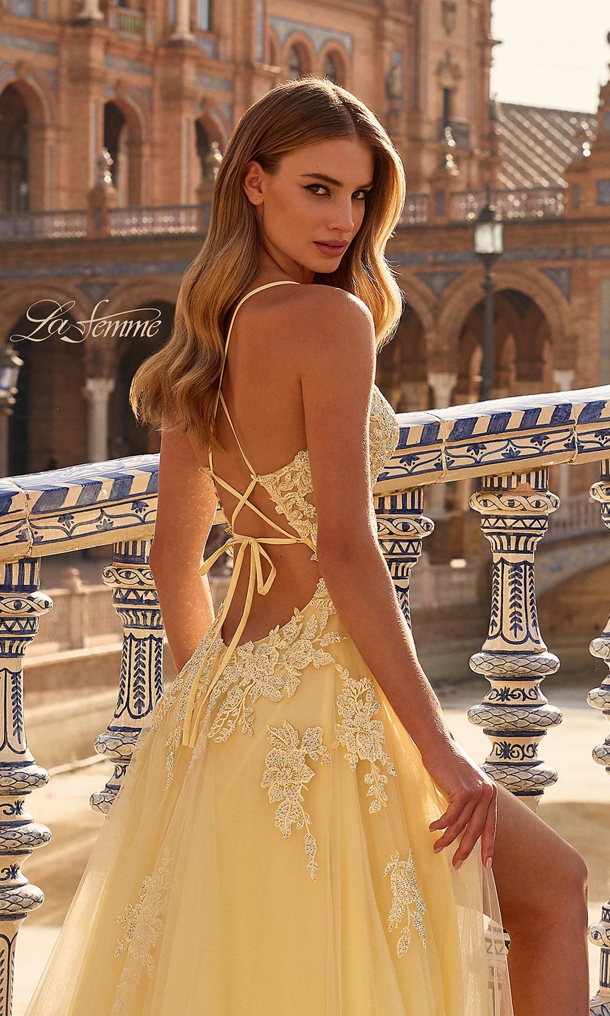 A woman in the La Femme 33716 long prom dress with pale yellow lace embroidery and a lace-up back stands by ornate railings, her tulle skirt flowing gracefully with historic architecture in the background.